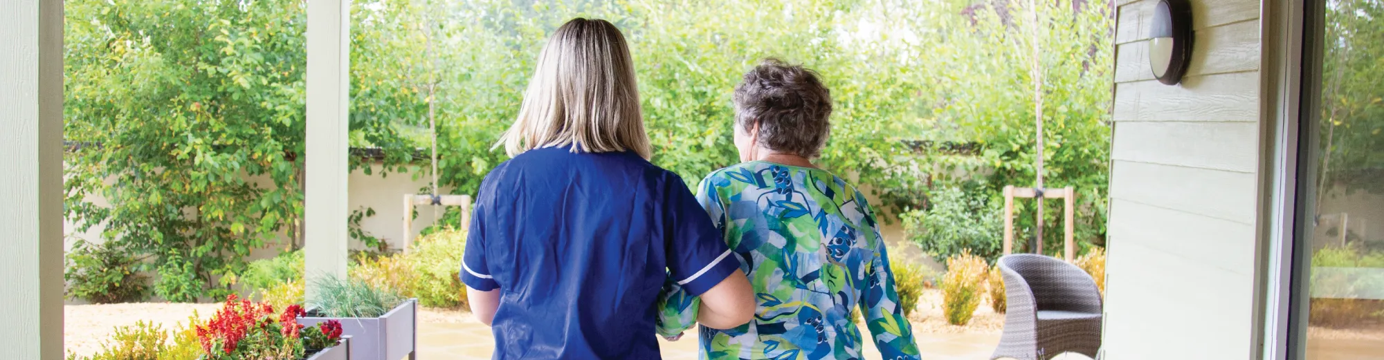 A carer in a blue uniform walking arm in arm with an older woman out onto a garden patio