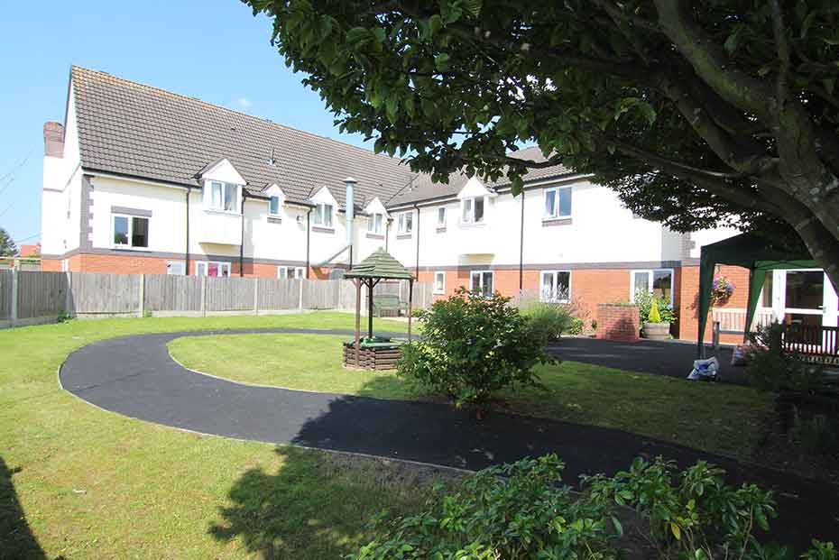 A two-story white building and garden area with a neatly trimmed lawn, a gazebo, and a paved path under a clear sky.