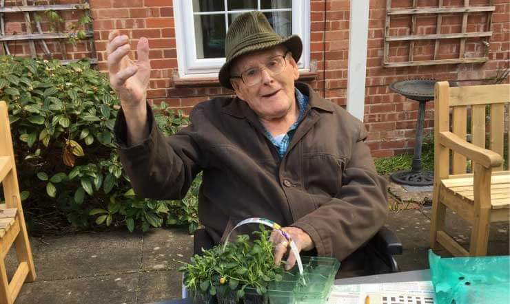 Elderly man raises hand whilst tending to plants at an outdoor table