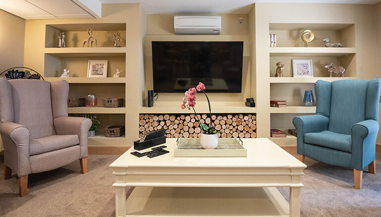 A well-decorated living room featuring a blue armchair and a pink armchair, a white coffee table, and a television above a decorative fireplace, surrounded by shelves with various ornaments