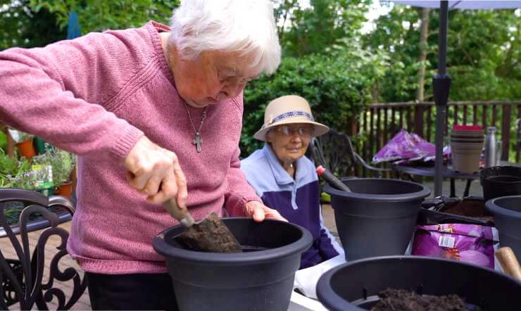 An elderly lady in a pink jumper gardening with a trowel and a black pot, with various gardening supplies on a table