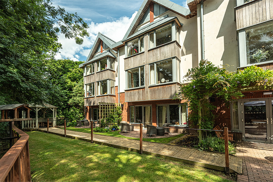 Multi-story  building with a combination of wooden and white exteriors, surrounded by lush greenery and a well-maintained and landscaped garden