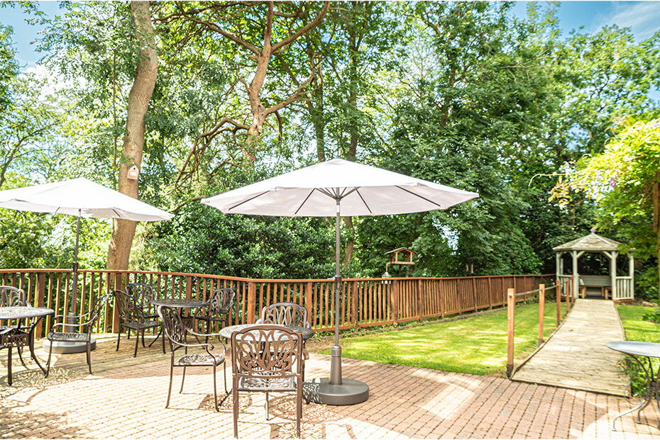 Outdoor patio area with multiple tables and chairs under white umbrellas, surrounded by green trees and a wooden fence