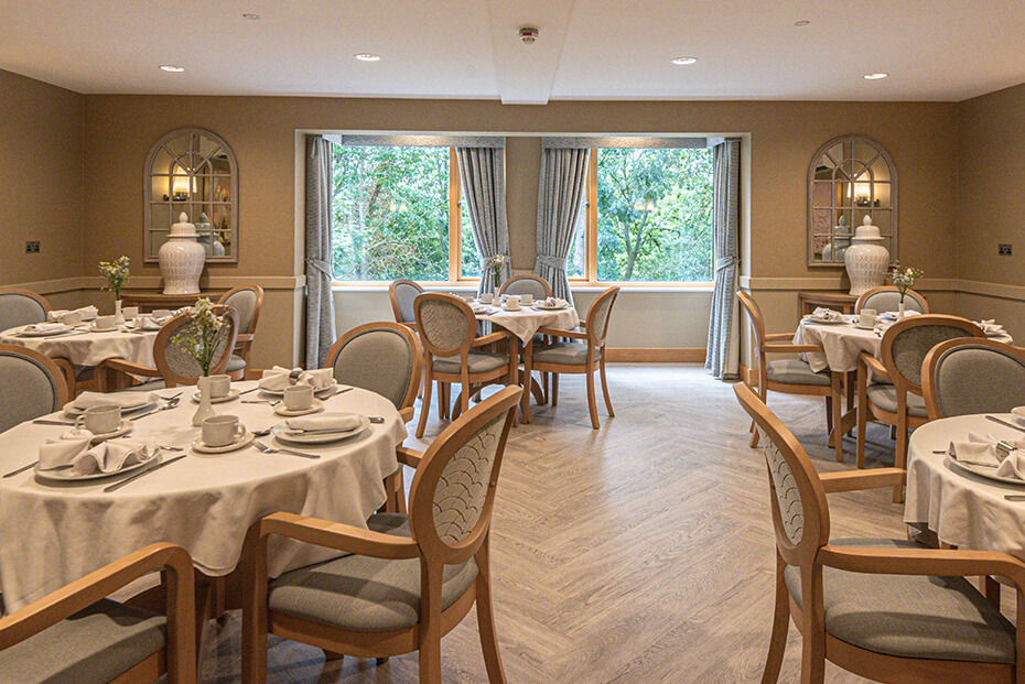 An elegant dining room with set tables, chairs, and a view of trees through large windows.