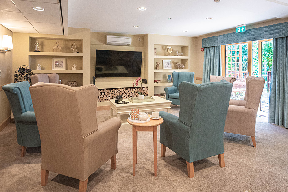 Inviting living room with a fireplace, a flat-screen TV, blue and beige armchairs, and a small round table with a teapot set, opening to an outdoor area through large patio doors.