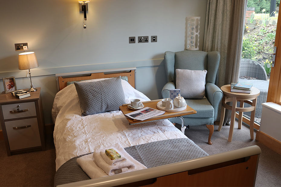 Cosy bedroom with a made bed, a bedside table with a lamp, and a sitting area with an armchair and small table set with tea service.