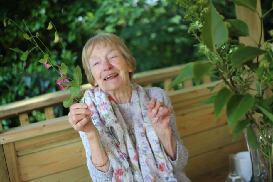 An older lady holding a flower in front of their face, sitting on a bench with green foliage in the background