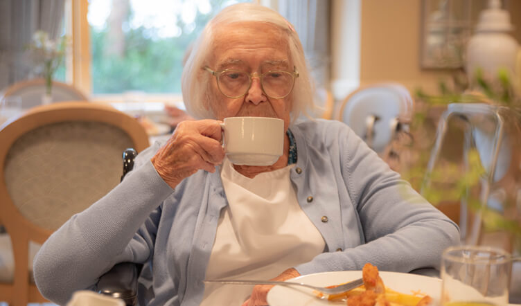 An elderly lady with white hair, wearing a blue cardigan, sits at a table in a dining room and sips of cup of tea