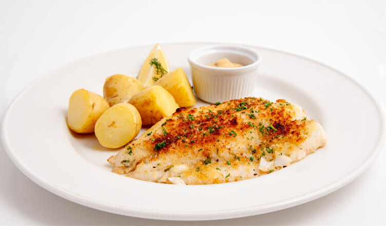 A plate of grilled fish fillet garnished with herbs, served with sliced boiled potatoes, lemon wedges, and a small bowl of sauce