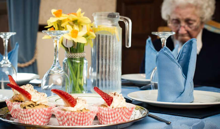 A well-set dining table with blue napkins, plates, and a pitcher of water, alongside with a plate of cupcakes