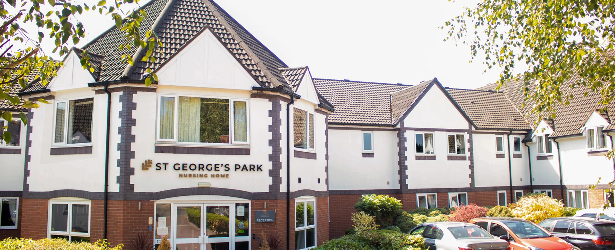 The front of St George's Park nursing home, a white and red brick building with tiled gables, a glazed reception entrance, and planting and parked cars to the front.