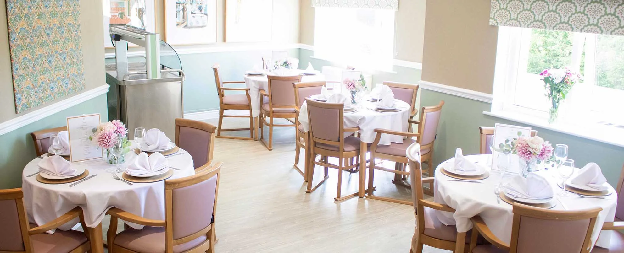 A bright dining room with sage green walls, round tables laid with white linen, napkins and glassware, pink upholstered oak chairs, and fresh flowers on every table.
