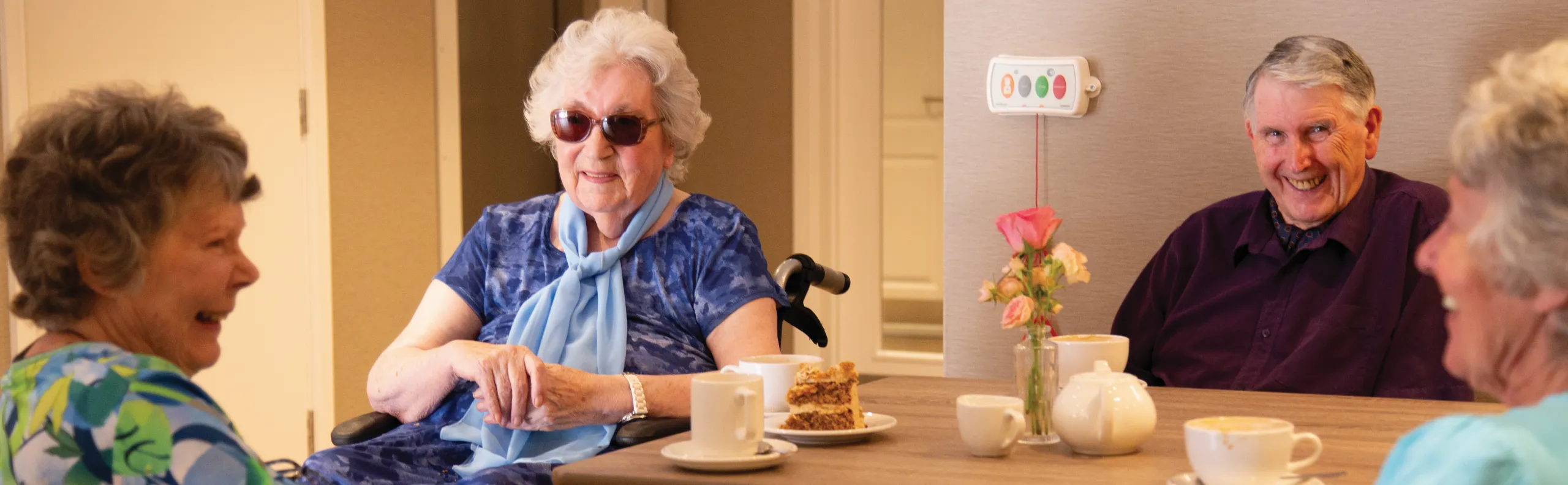 Carole, in sunglasses and a pale blue scarf, laughing with Dennis and two other women over tea and cake around a table
