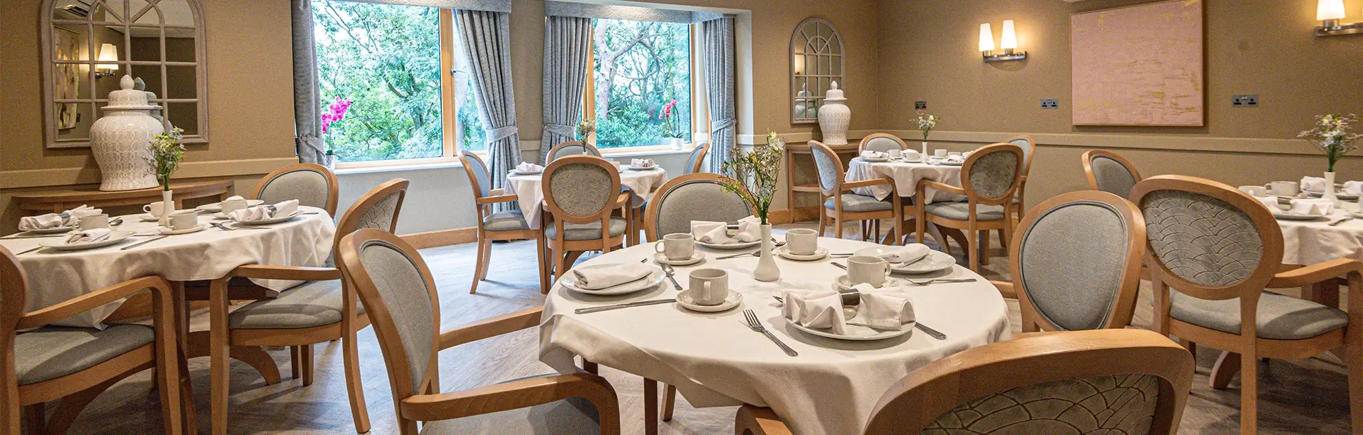 An elegant dining room with set tables, chairs, and a view of trees through large windows.