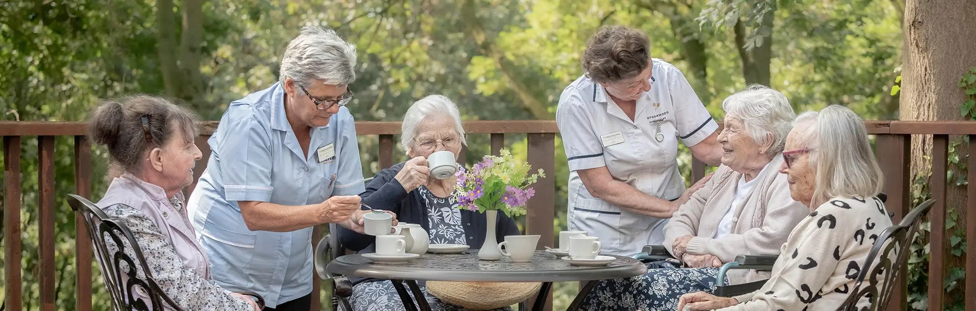 A group of elderly ladies sitting around a table outdoors with healthcare workers, enjoying a tea party.