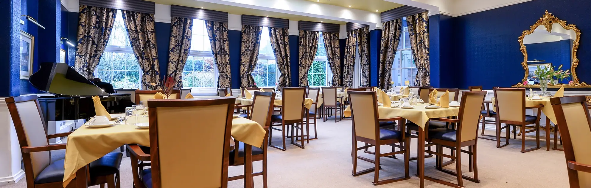 An elegant dining room with multiple set tables, beige chairs, blue walls, and large windows with patterned curtains.