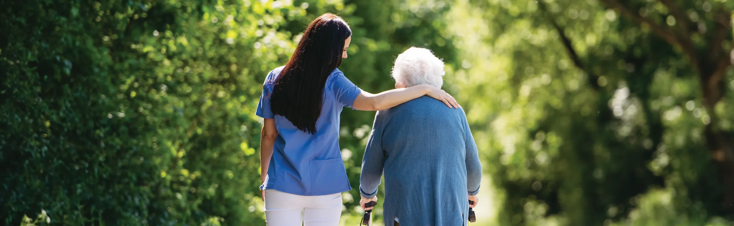 A carer in blue scrubs with her arm around the shoulders of an older woman using a walking aid, walking along a tree-lined path