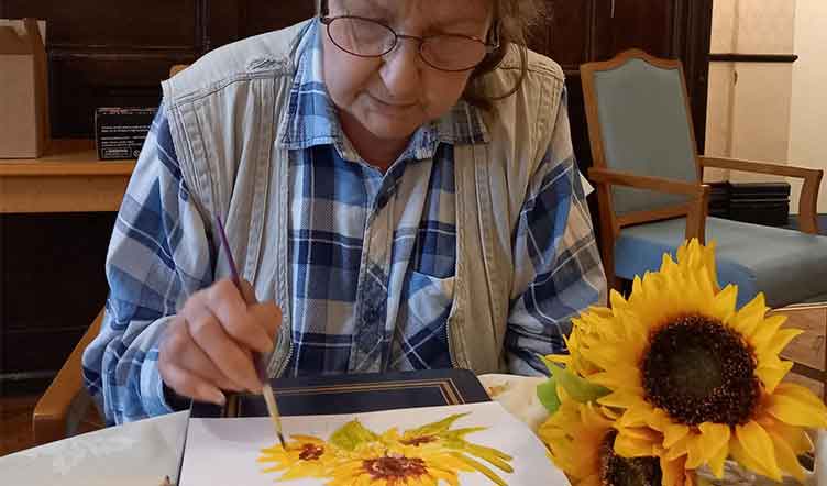 An older lady painting a sunflower on a canvas, with a real sunflower beside for reference