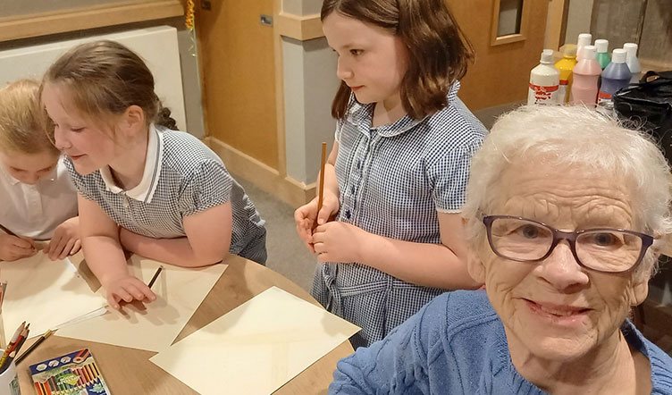 A group of children and an elderly woman engaged in an art activity. The children are drawing and colouring on paper, and the elderly woman is smiling at the camera
