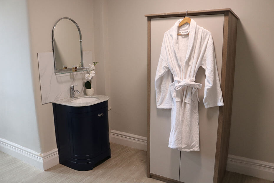 A bathroom corner featuring a dark vanity sink with a small vase with flowers on and a white robe hanging on a wooden wardrobe.