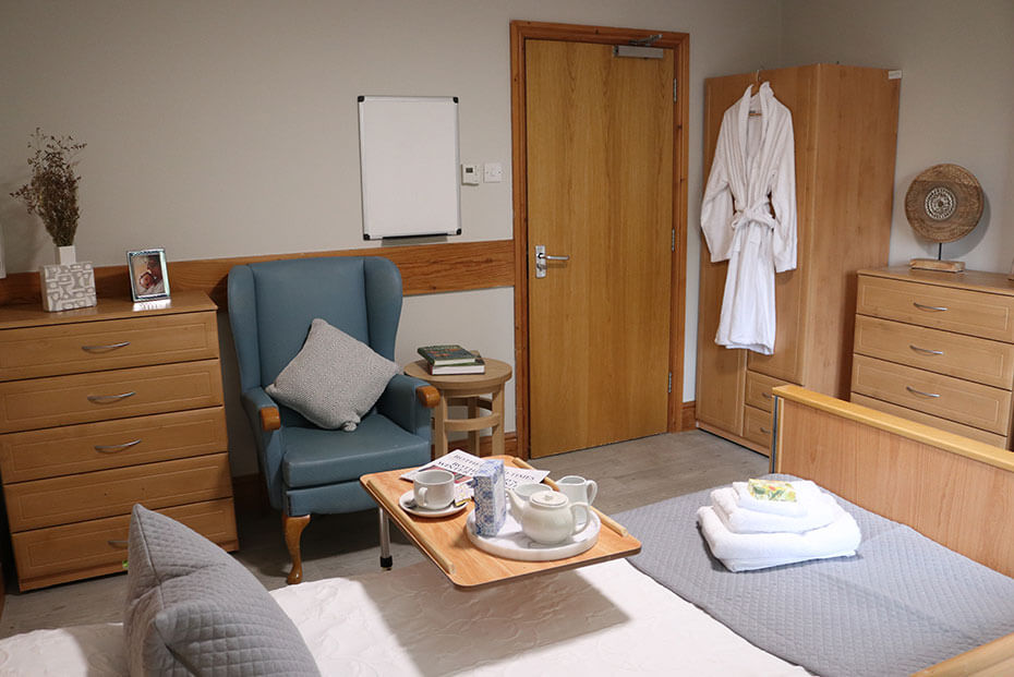 A cosy bedroom with a blue armchair, a small wooden coffee table with a tea set and wooden furniture including drawers and a wardrobe