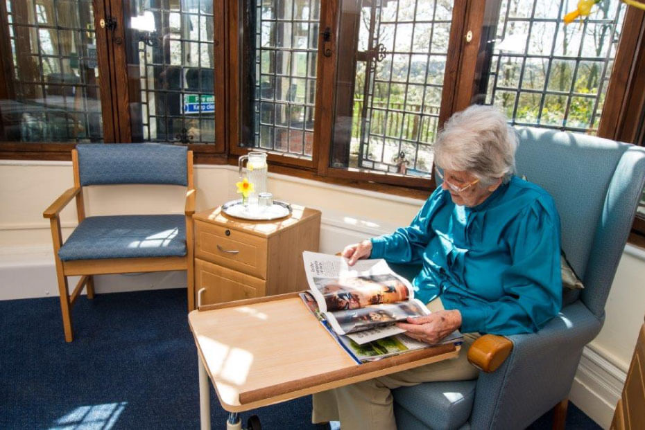 An elderly lady reading a magazine, seated comfortably in a sunlit lounge with large windows.