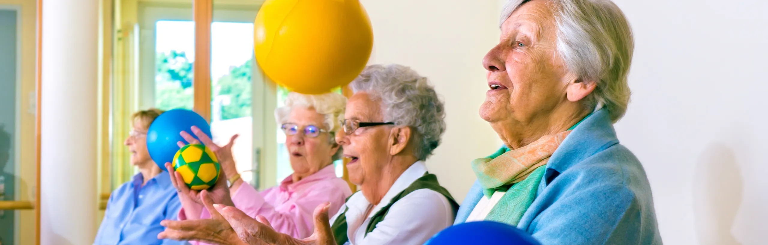 A group of older women taking part in a seated exercise class, passing coloured balls between them