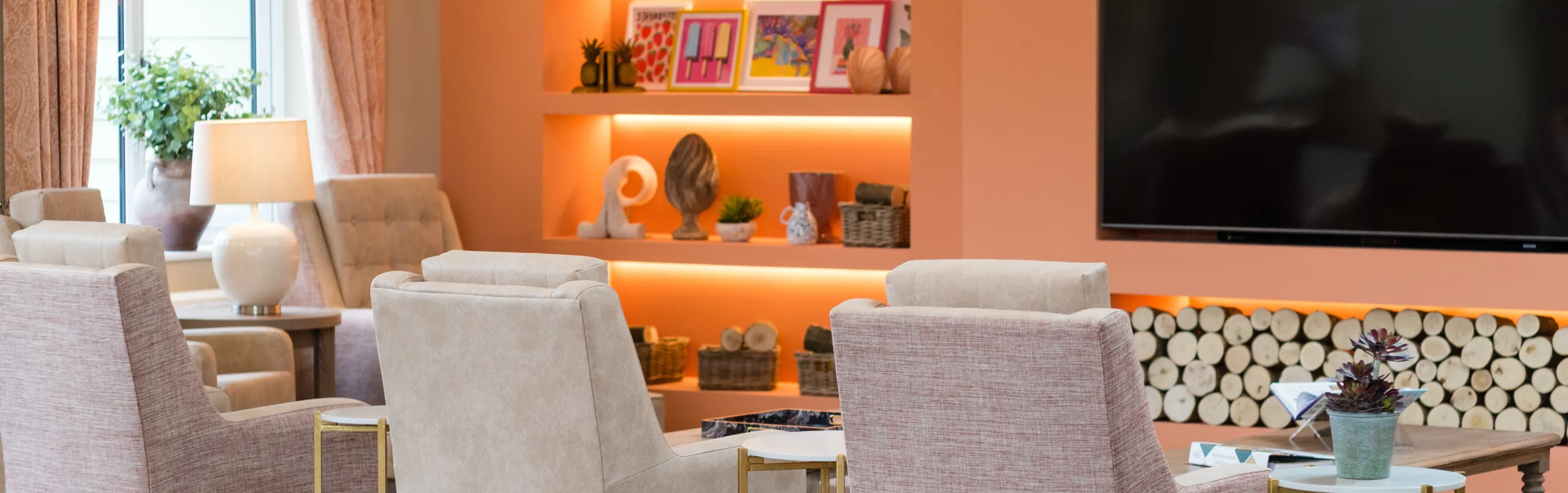 A lounge at Colwall care home with armchairs, a lit alcove of ornaments and a wall-mounted television