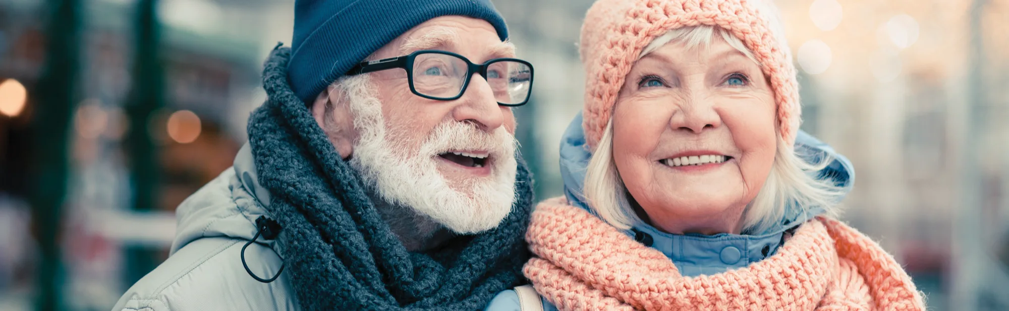 An older couple wrapped up in woolly hats and scarves, smiling together outdoors on a winter’s day