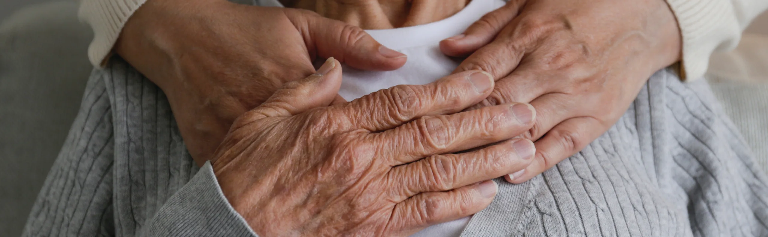 A carer’s hands resting gently over the clasped hands of an older person in a grey cardigan