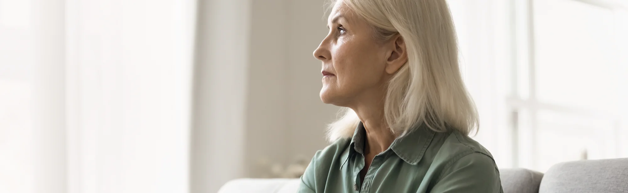 An older woman with white hair sitting beside a bright window, looking thoughtfully into the distance