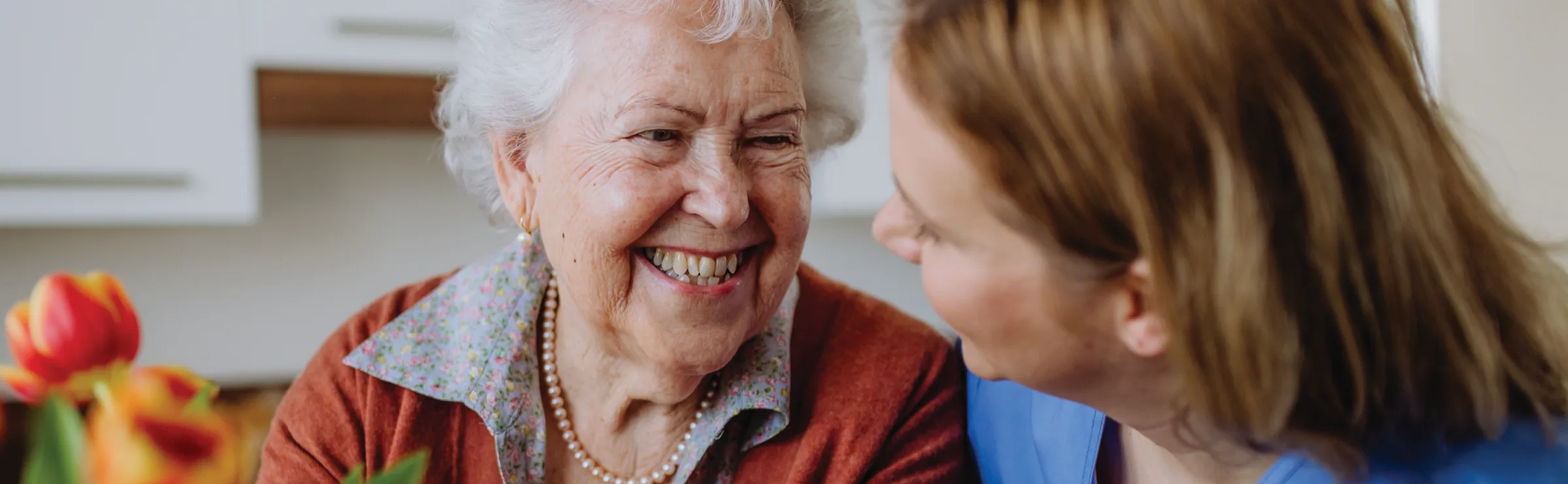 A smiling older woman with white hair face to face with a carer, with red and orange tulips in the foreground