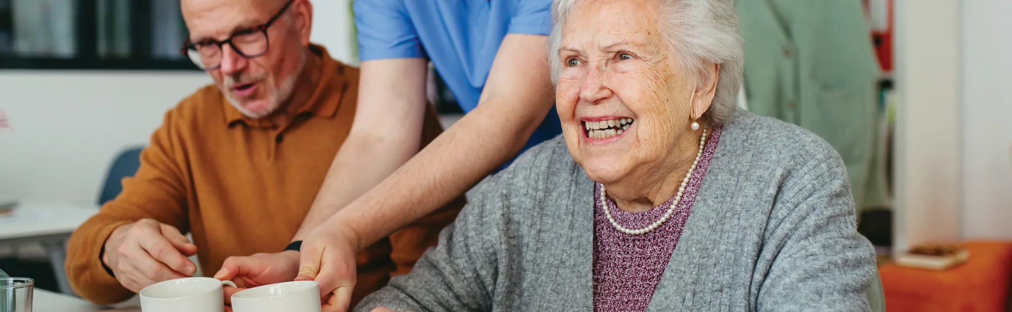 An older woman laughing at a table beside a carer, with a man in an orange jumper stirring a cup of tea