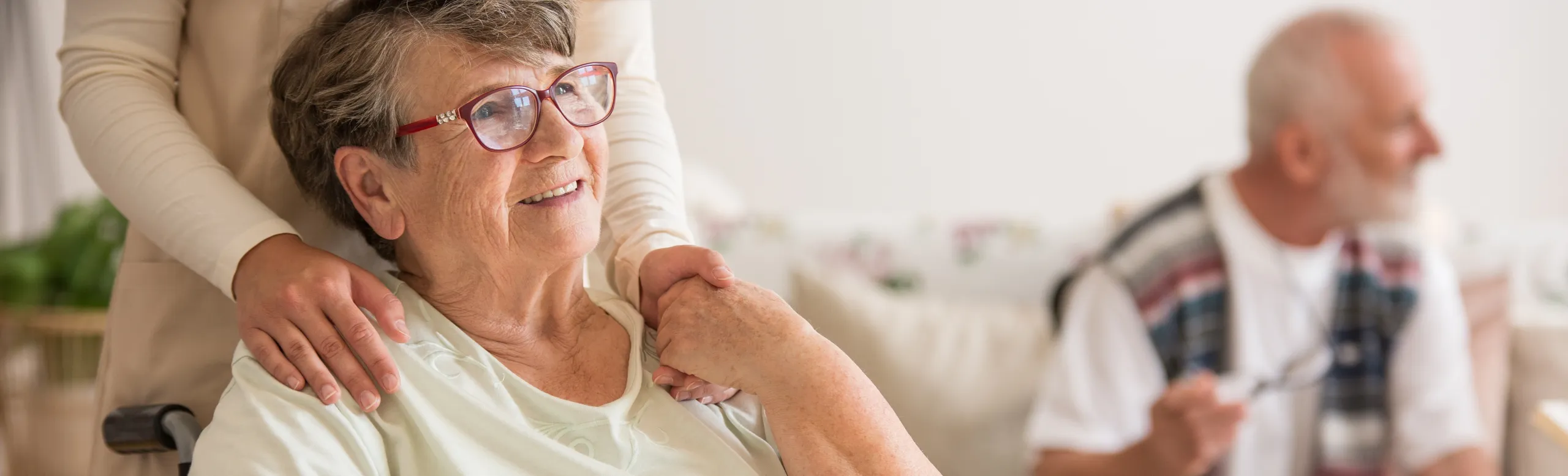 A carer resting a hand on the shoulder of a smiling older woman in a care home lounge