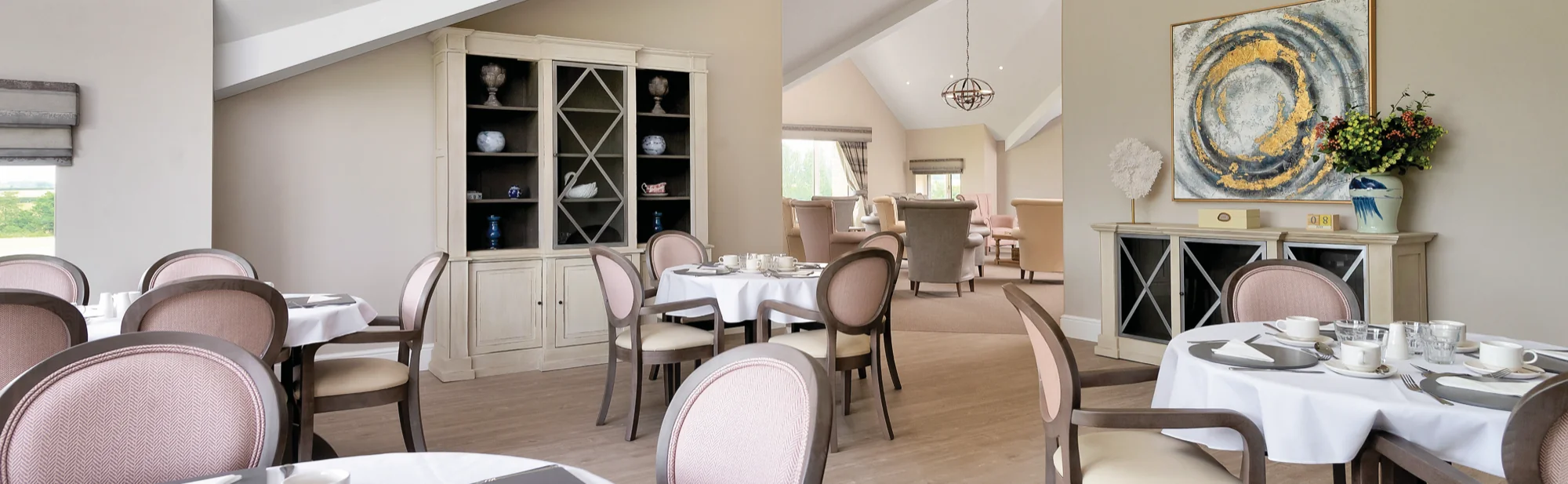 A care home dining room with linen-laid tables, dusky pink chairs and a display cabinet
