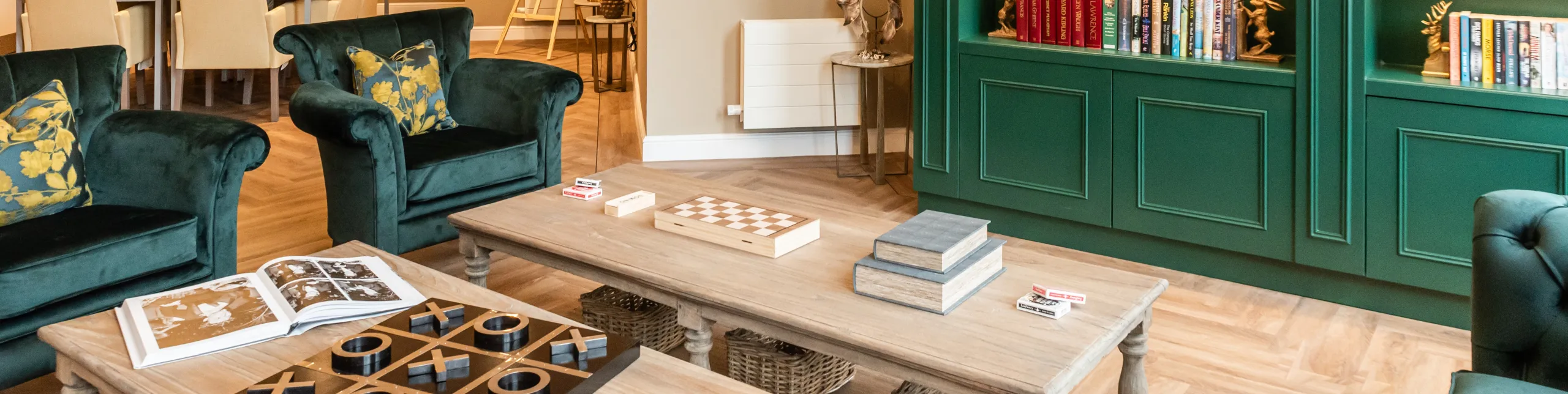 A lounge at Colwall care home with velvet armchairs, board games on low tables and a green bookcase