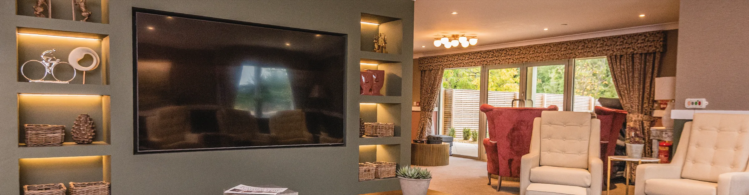 A care home lounge at Colwall with a television set into lit display shelving, and red and cream armchairs by the garden doors