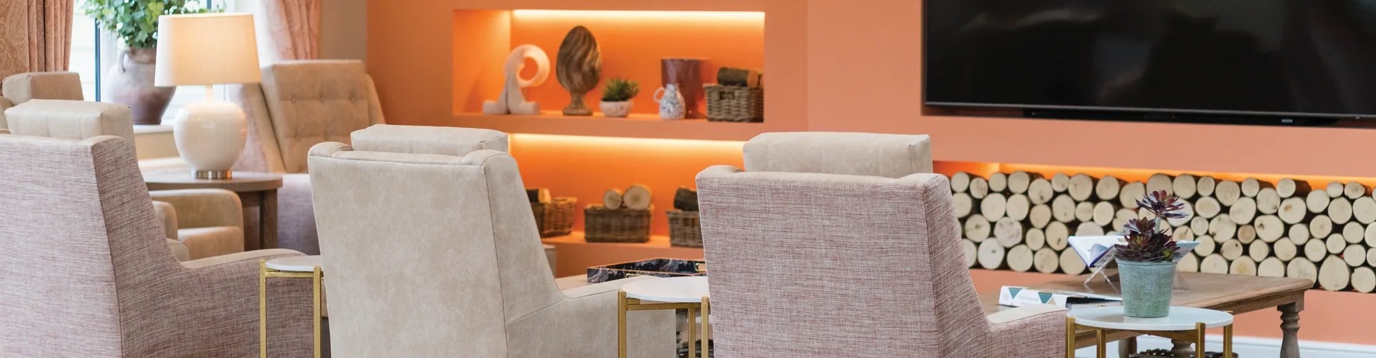 A care home lounge with peach walls, a lit display alcove, a wall-mounted television and pink armchairs