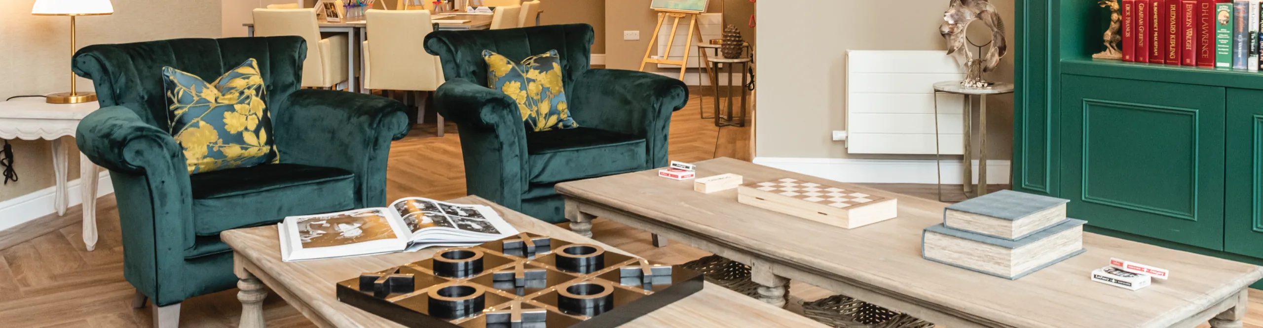 A care home lounge with green velvet armchairs, low tables set with board games and a bookcase