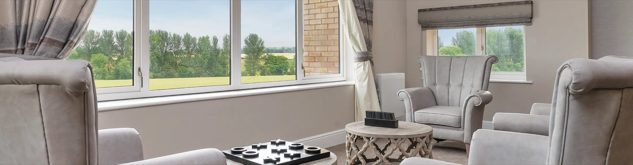 Grey armchairs around a low table in a care home lounge, with wide windows looking out over fields and trees