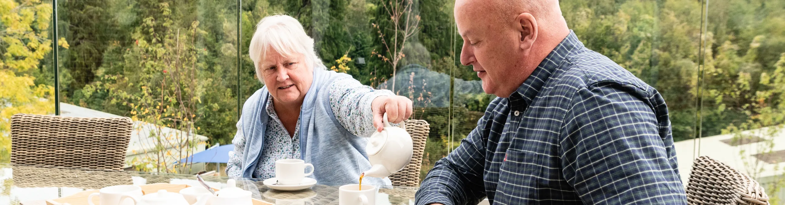An older woman pouring tea for a man at an outdoor table overlooking woodland