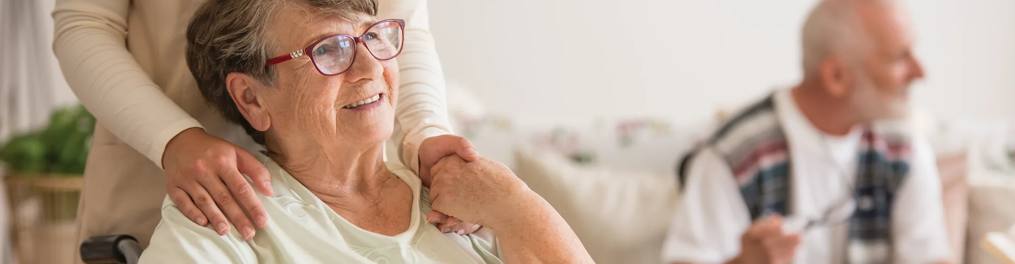 A carer resting both hands on the shoulders of a smiling older woman wearing red glasses
