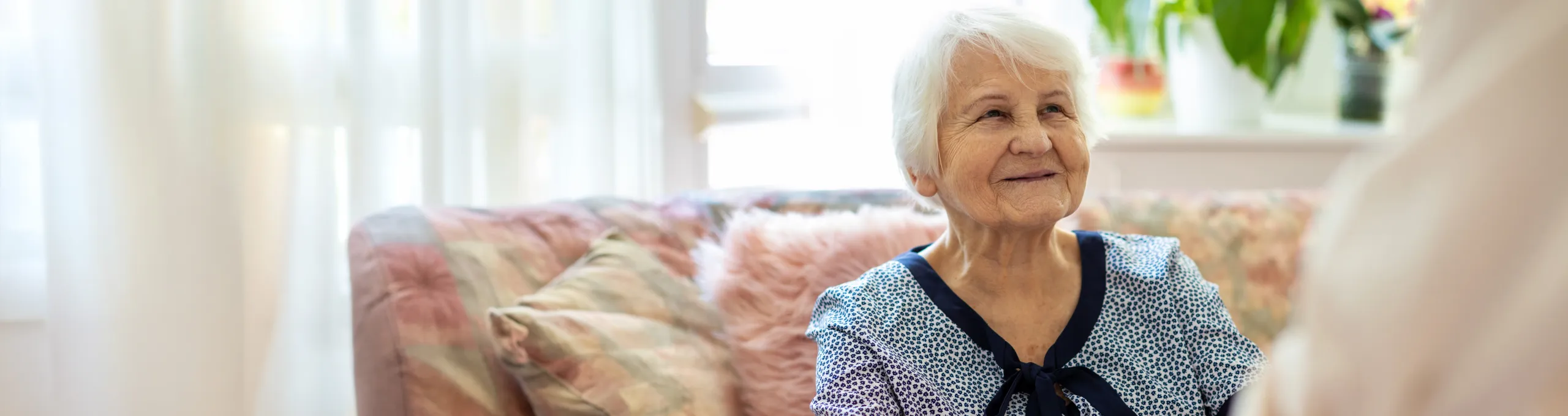 An elderly woman sitting on a floral sofa in a bright room, looking towards the window