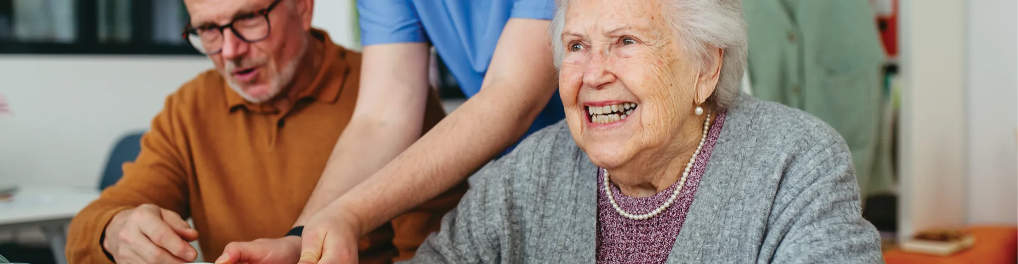 A smiling older woman in a grey cardigan and pearls sitting at a table beside an older man, with a carer leaning in between them