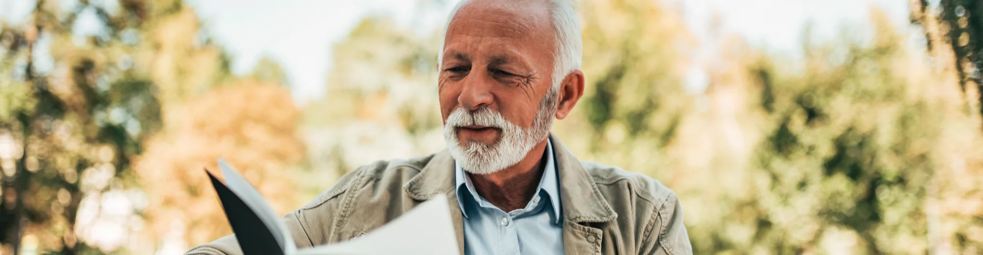 An older man with a white beard reading a book outdoors, surrounded by autumn trees