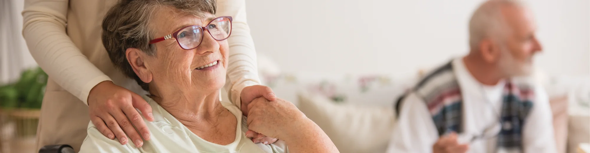 A carer resting both hands on the shoulders of a smiling older woman wearing red glasses