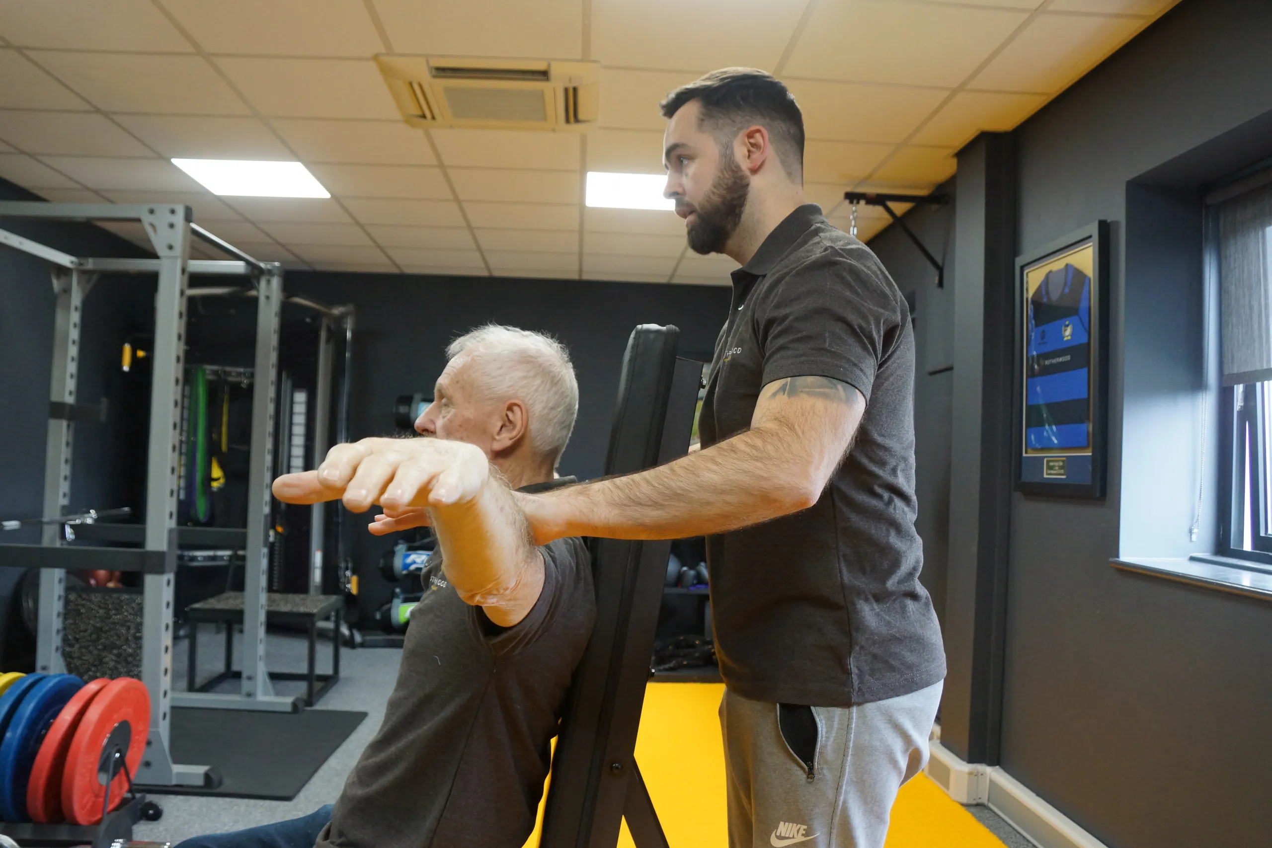 A wellness coach guiding an older man through a seated arm exercise in a gym