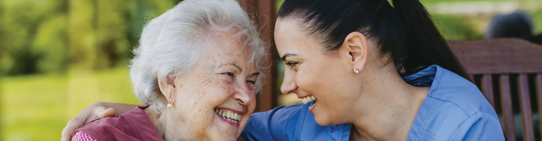 An older woman laughing with a carer in blue scrubs who has an arm around her shoulders