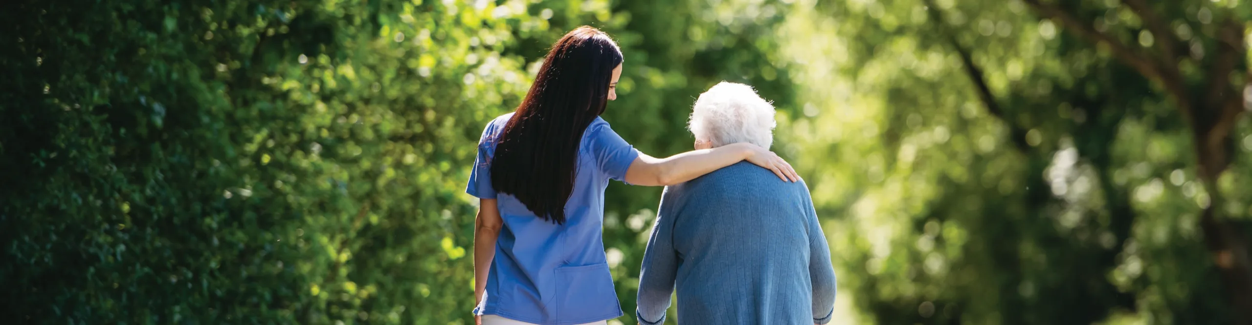 A carer in blue scrubs walking outdoors with an older woman, an arm around her shoulders