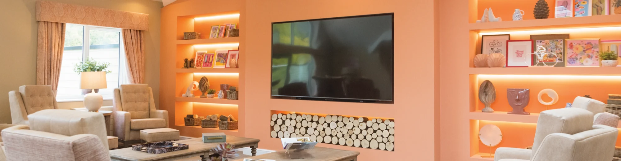A care home lounge with coral walls, lit display alcoves, a wall-mounted television and armchairs